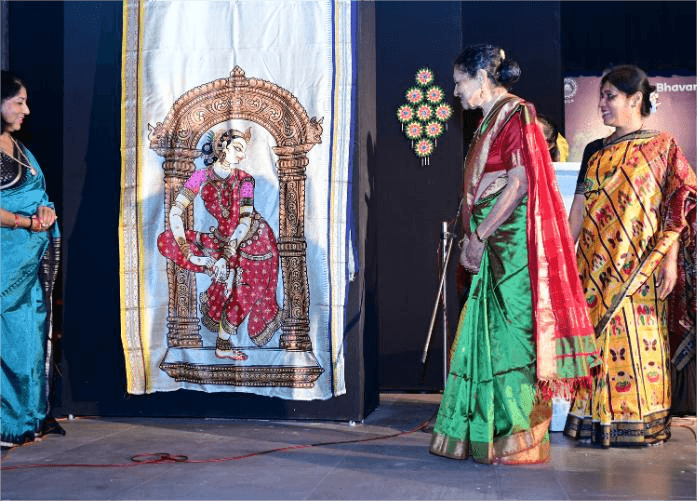 Three women on stage, central element is large vertical fabric panel featuring detailed painting of dancing figure in traditional attire framed by archway, two women in sarees standing on either side of panel observing or facing audience