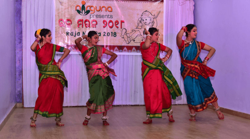 Four women in traditional Indian dance costumes (sarees in shades of red, green, and blue) performing on stage in various dynamic dance poses, white banner with text in Indian script and 'Raja N 2018' visible in background